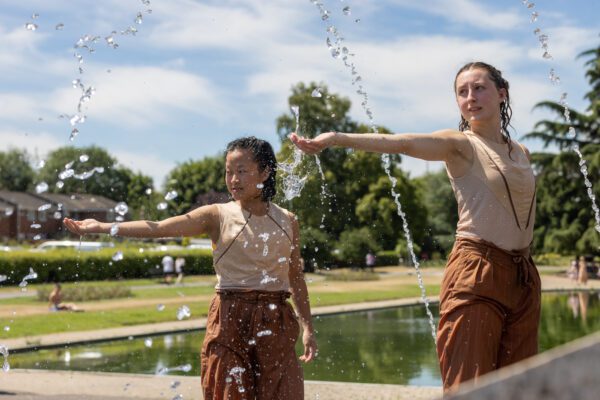 Two performers reaching out to the audience with a water feature in the background and spouting water in the foreground.