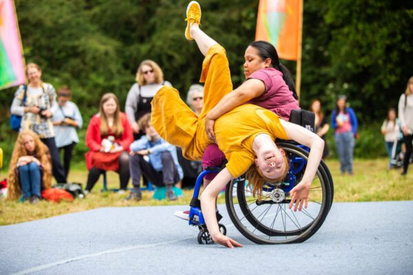 One person sat on a wheelchair, another person is leant over them with their legs in the air and hands reaching for the floor. Audience members watch in the background.