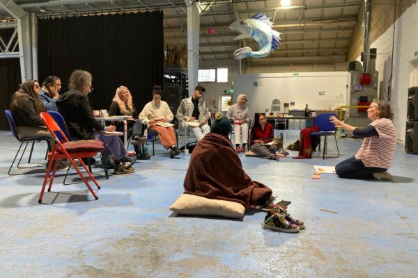 A group of people sat in a warehouse on chairs and the floor. There is an instructor at the front who is gesturing to the group.