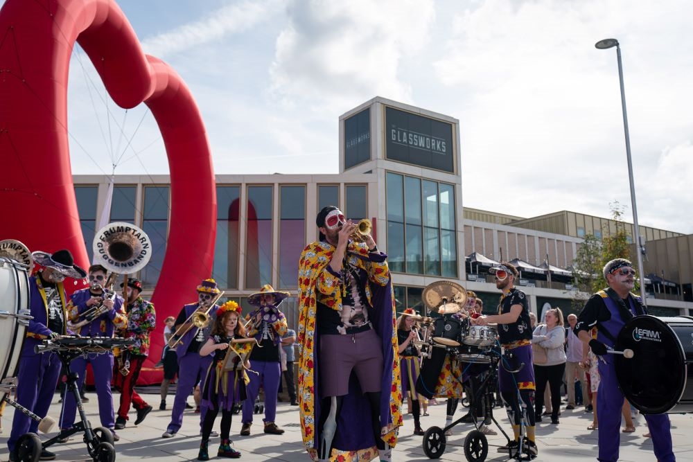 A brass band with skeletal bodysuits on under purple clothing and skulls painted on their faces play their instruments outside the Glassworks building in Barnsley.