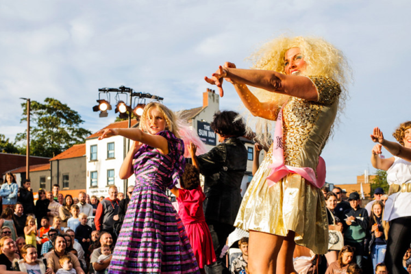 A wide angle shot of the Eau de Memoire performers all pointing just off-centre of the direction of the camera. Miss High Leg Kick wears a large, blonde, permed wig and a gold sparkly dress. All performers wear an array of colourful 80s outfits and wigs. Without Walls TALKS: Creating a Sustainable Production by Francesca Baglione