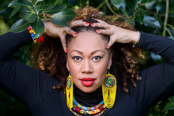 A portrait image of a black woman with her hands on her head. She is wearing long yellow decorative earrings, a decorative necklace and a black top.