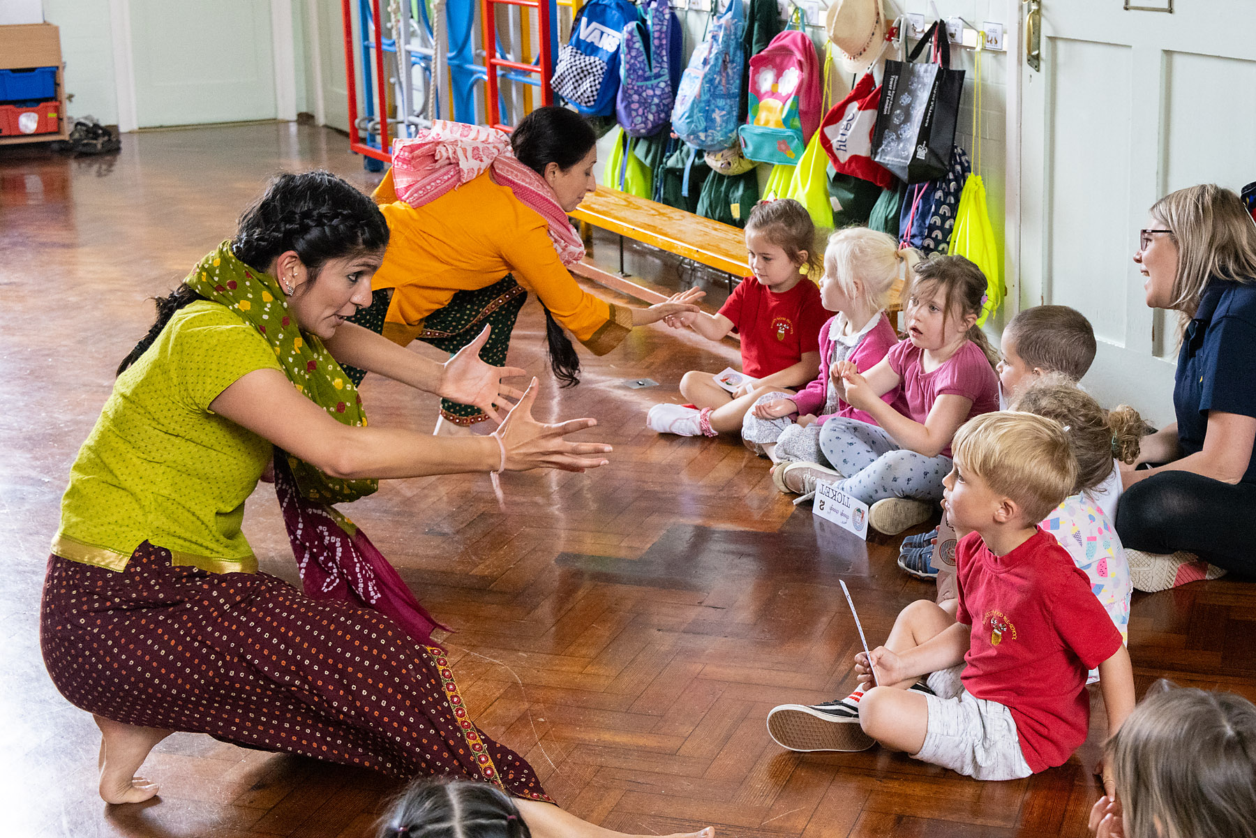 Two women of South Asian heritage are dressed in traditional South Asian clothing. They are knelt down on the floor in front of a row of school children. Their hands point towards the children as the children gae back at the performers.