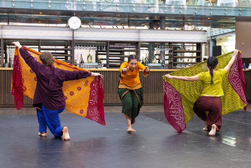 3 performers weraing traditional South Asian clothing. Two performers kneel on the floor holding up scarves facing away from the camera. The scarves are angled toward the middle where another performer runs through. Their arms are pointed forward and their head tucked bewteen them.