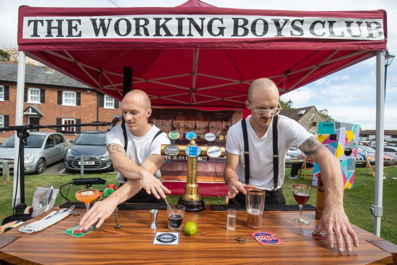 Two white men with shaved heads wearing white t-shirts and braces over the top. The are stood behind a bar playing the bar DJ set. They are under a red canopy that says 'Working Boys Club'.