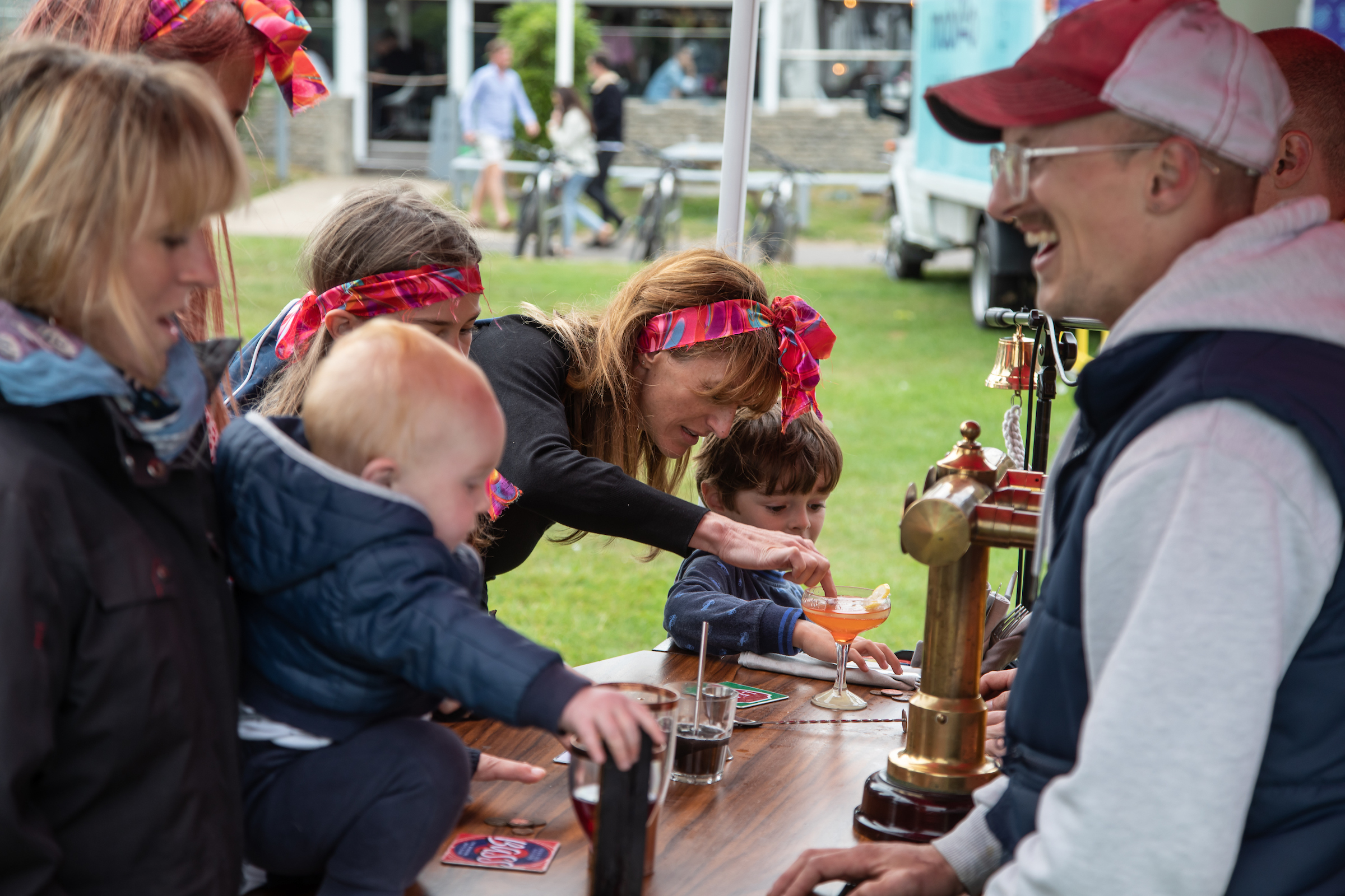A group of people with small children all lean over the bar to try out the DJ-ing. One of the Working Boys is wearing a red and white cap with a grey hoodie and navy gilet. He is similing.