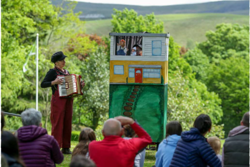 A person dressed in navy and red stands next to a puppet booth playing an accordian.