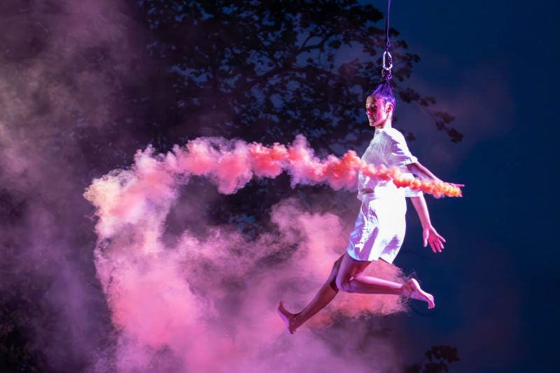 An image of a circus performer suspended in the air with a horizontal pillar of pink smoke in front of her with a black background,