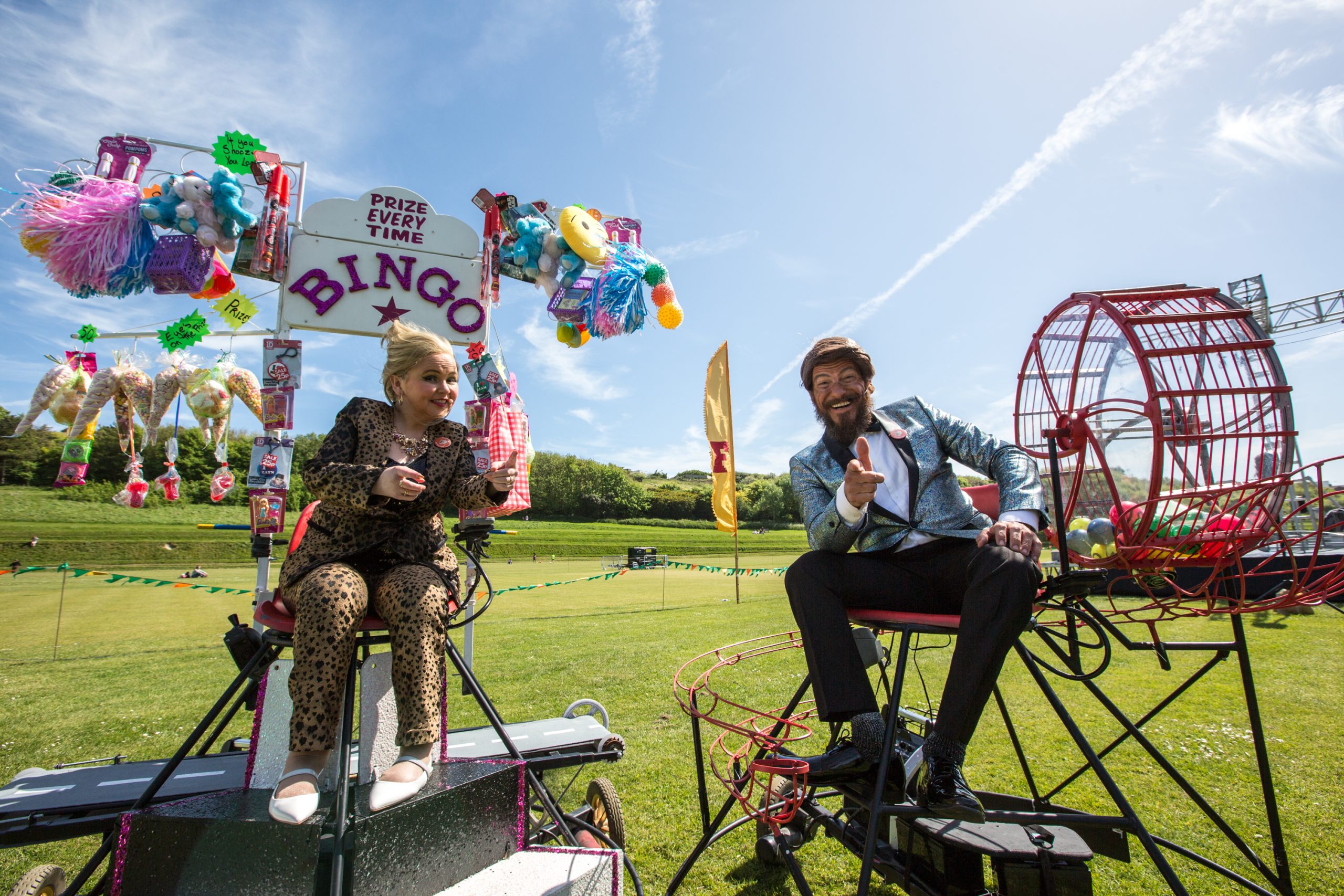 An image of a male and female performer sat on two different platforms in a field. There are fairground props around them and a blue sky background.