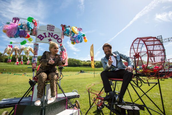 A white woman and white man sit on a set bingo set in a field. The woman has blonde hair and wears a leopard print outfit. She sits under a sign that says 'Bingo' and has lots of colorful toys and products hanging down from it. The man wears a blue blazer and black trousers. He is sat next to a bingo cage.