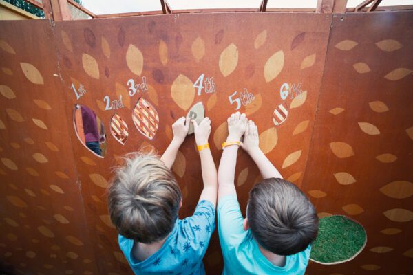 An image of two children with their backs to the camera pinning an object onto a makeshift orange wall with a decorative leafy pattern and numbers 1 to 5.