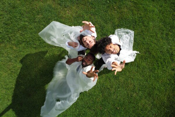 This photo is taken from above three performers stood with their backs together in a small circle. They are standing on green grass and covered in loose flowing white plastic. They are all looking up into the camera stretching an arm towards it. They all look scared. Without Walls TALKS: Theatre Témoin on integrating BSL