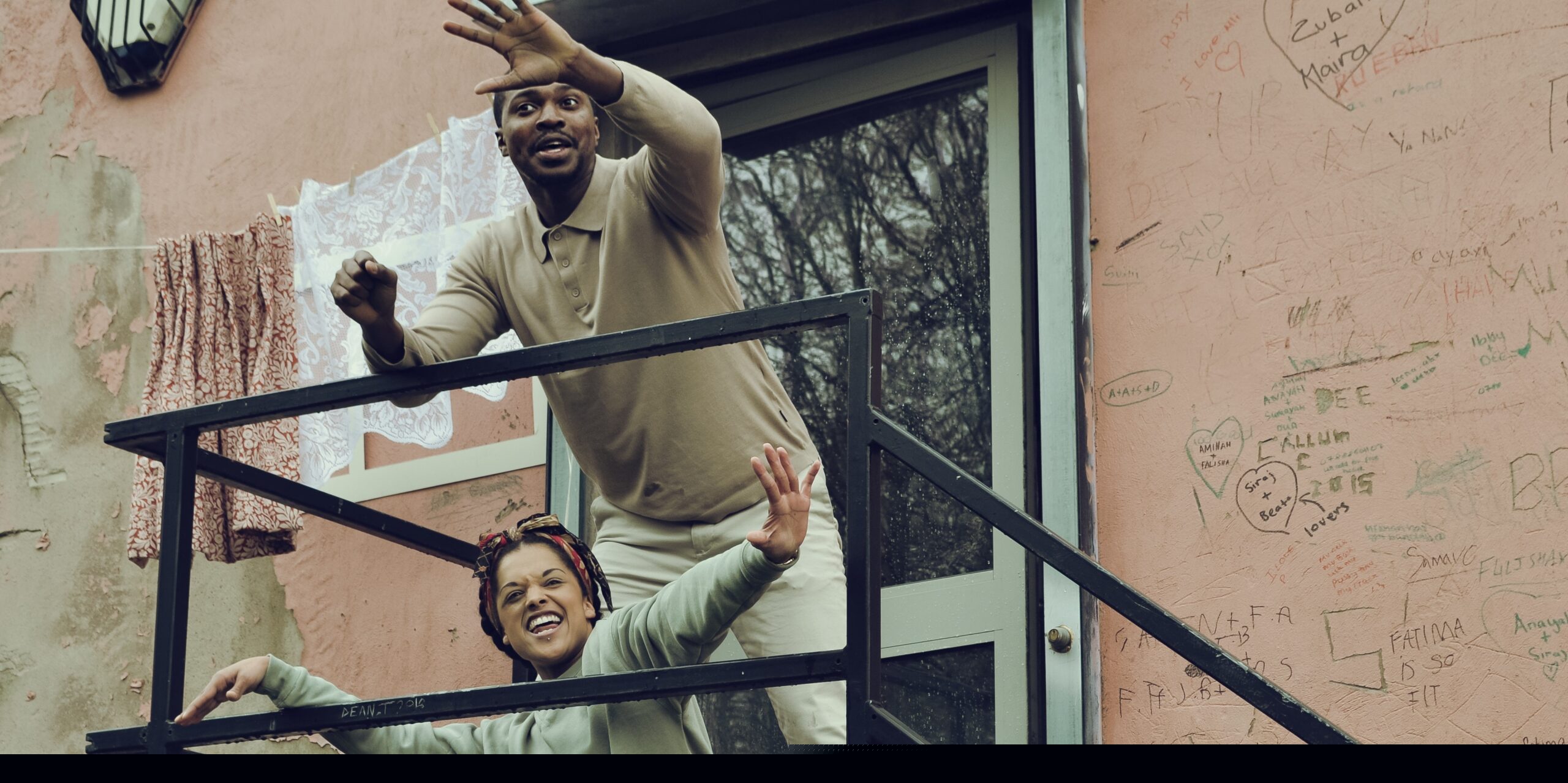 Two performers are at the top of the stairs to a front door. One is standing behind the other who is sat down. They're both smiling and have their arms outstretched. There is laundry on the line to their right and pen graffiti on the wall to their left.