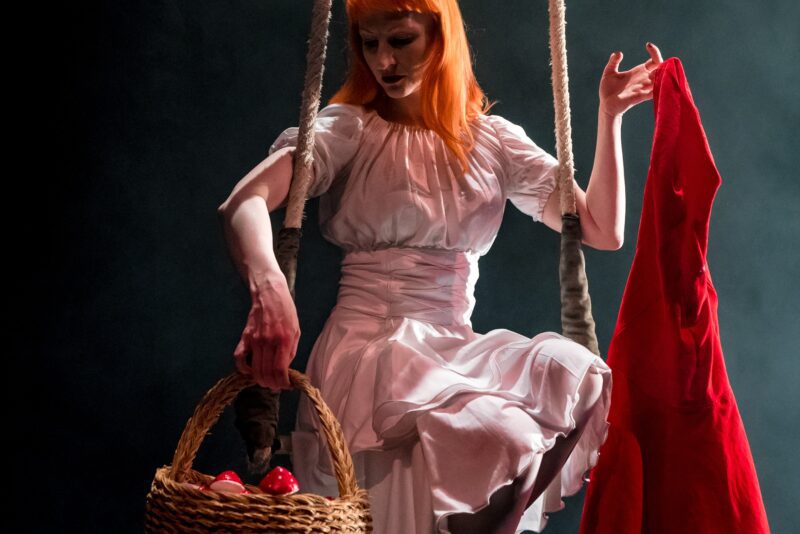 Vee Smith is sat on a trapeze with a white dress, holding a red cape with a red hood, holding a basket filled with red toadstools.