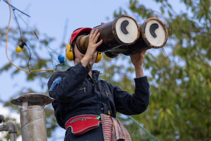 An image of a male performer looking through some giant binoculars. He is wearing noise cancelling headphones and a dark blue jacket.