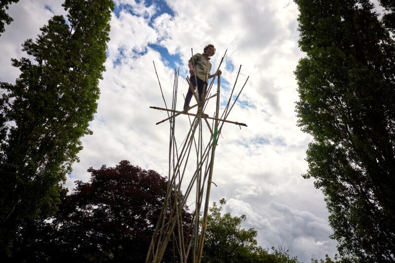 An image of a person suspended on wooden poles with a cloudy sky background with tall trees on both sides,