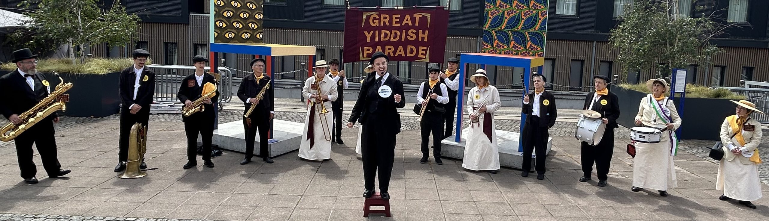 An image of a marching band stood in a row with a banner in the bakcground saying 'Grreat Yiddish Parade'