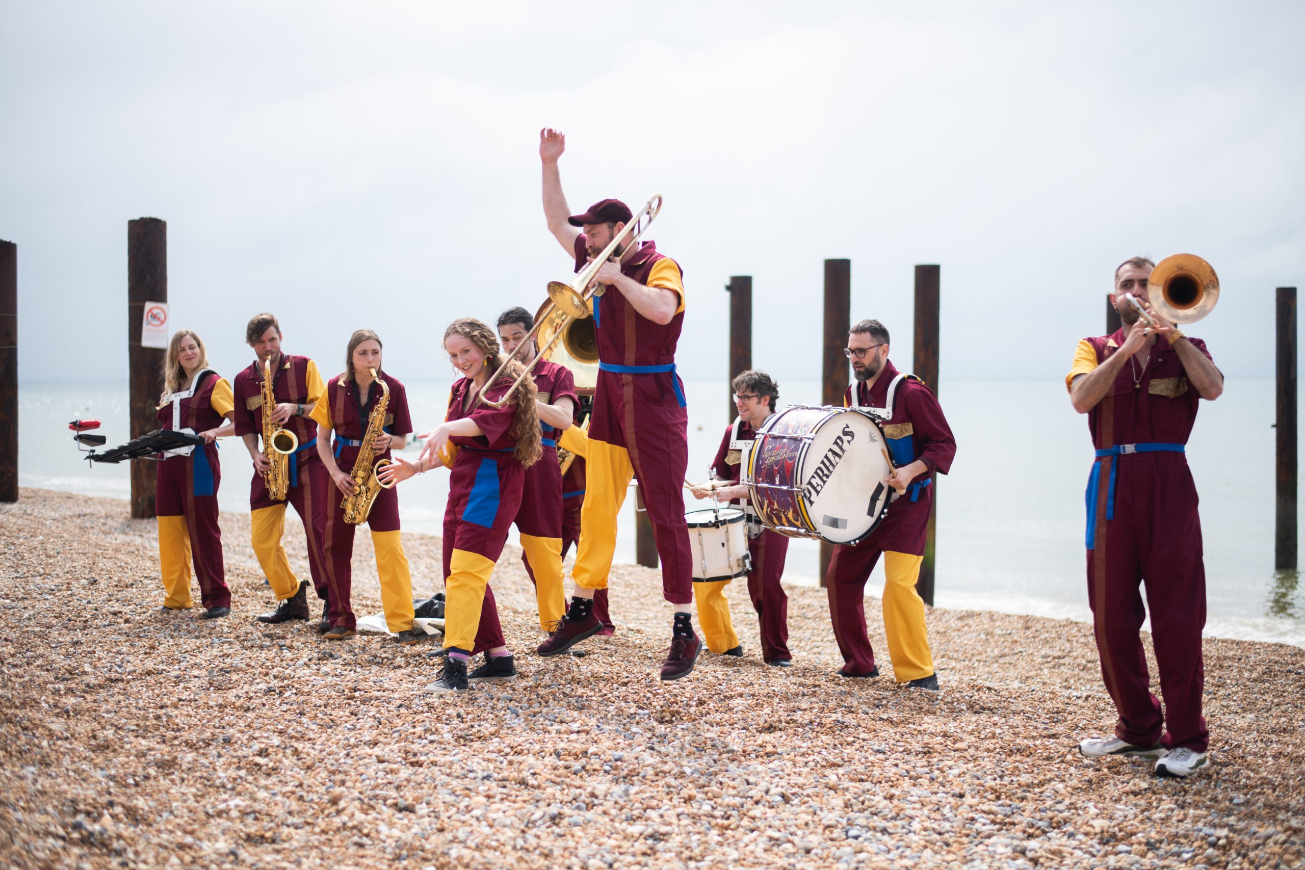 An image of a brass band stood on a beach playing various musical instruments. They are all wearing red and yellow clothing.