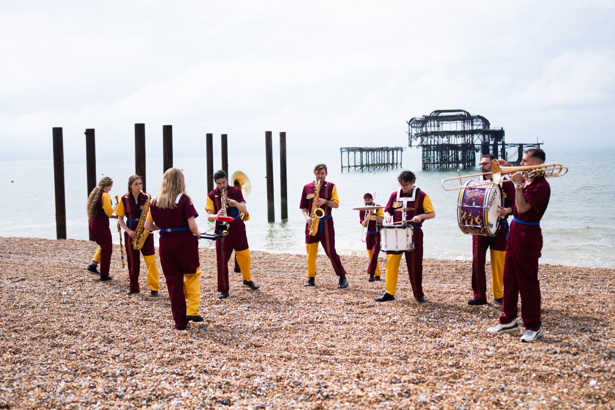 An image of a brass band stood on a beach playing various musical instruments. They are all wearing red and yellow clothing.