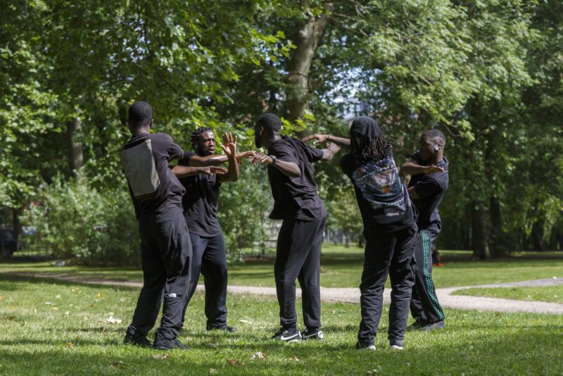 An image of a group of male performers dressed in black on a grassy area.