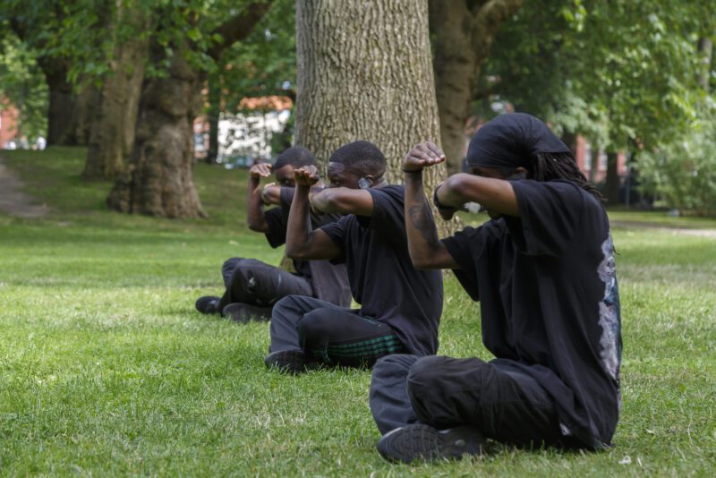 An image of a group of male performers dressed in black sat crossed legged on some grass mimicking arm gestures.