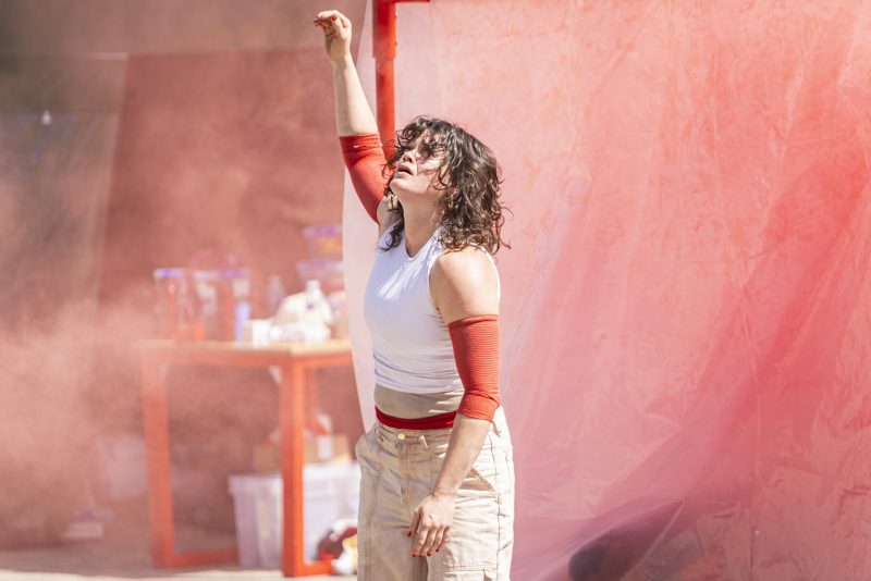 An image of a female dancer with her arm outstretched and a red sheet in the background.