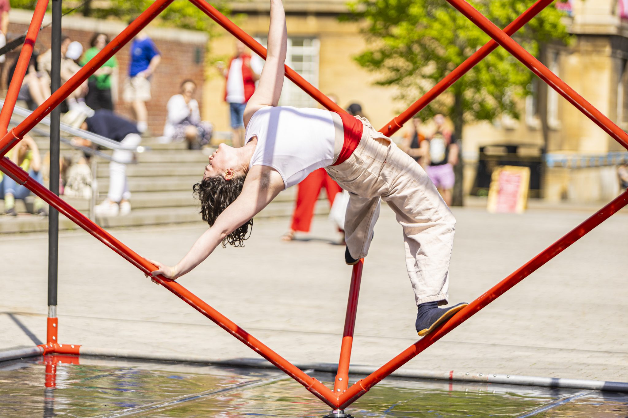 An image of a female performer suspended on a cube shaped frame who is leaning backwards. Sheis wearing a white top and cream trousers.