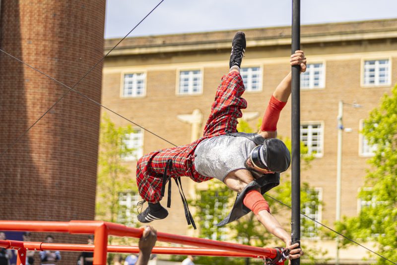 An image of a male performer suspended on a pole with a block of flats in the background. He is wearing checked trousers, a white vest and leather cap.
