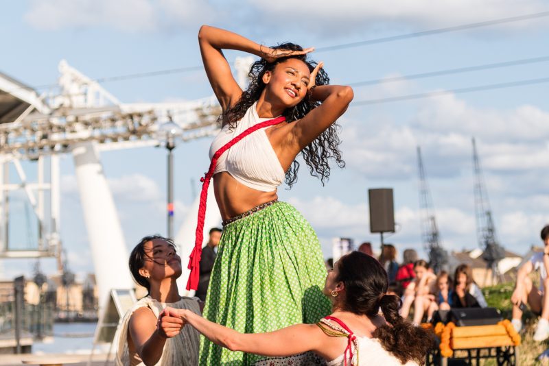 An image of a South Asian female dancer stood up with her arms over her head, with two female dancers sat around her in a circle and holding hands. She is wearing a white crop top and long green skirt.