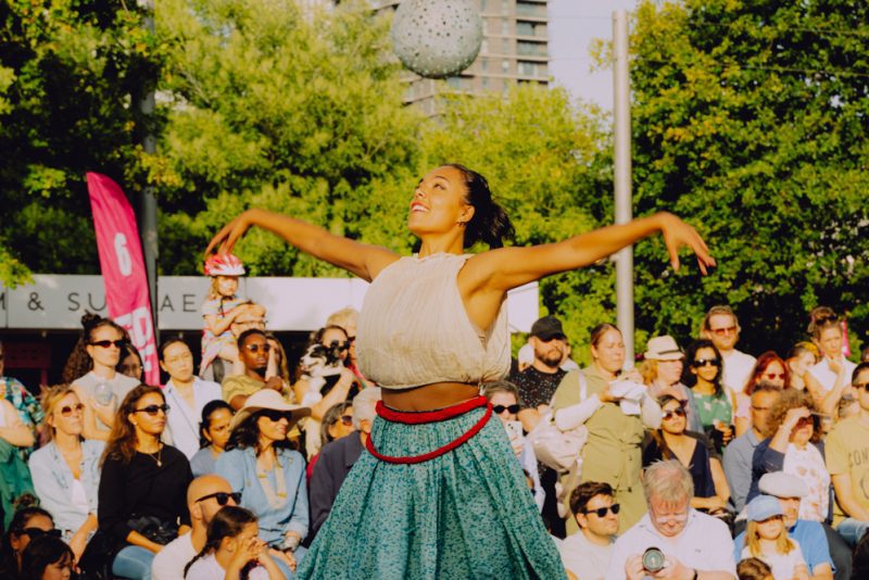 An image of a South Asian female dancer with her arms outstretched and a small audience behind her.