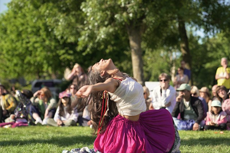 An image of a South Asian female dancer kneeling on a patch of grass with her head tilted backwards and her arms outstretched.