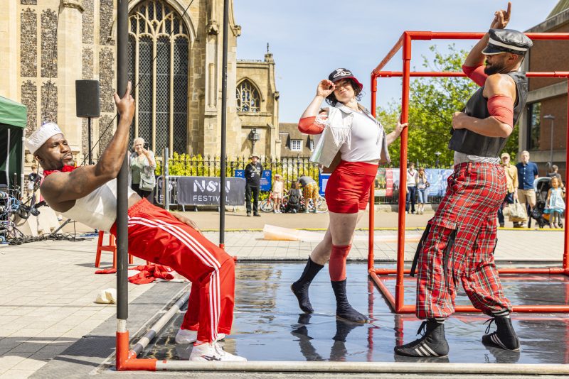 An image of three performers on a makeshift surface with a cube shaped frame and church in the background.