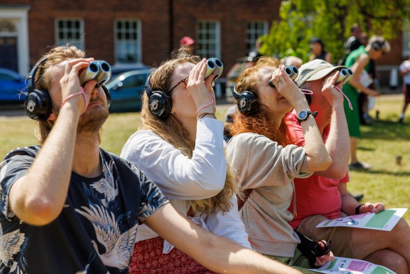 An image of four people sat down on some grass wearing headphones and looking through binoculars.