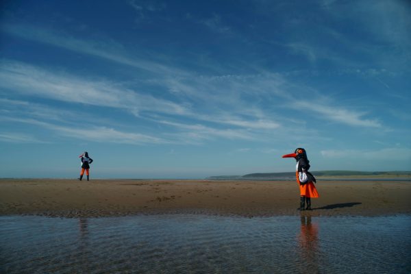 An image of a beach with a person dressed as a bird in the foreground and a figure in the background. The sky is blue with wispy clouds.