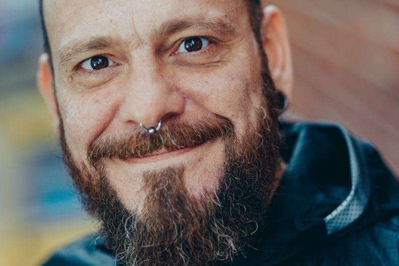 A portrait image of a man with a beard and a moustache and a nose ring wearing a blue top.