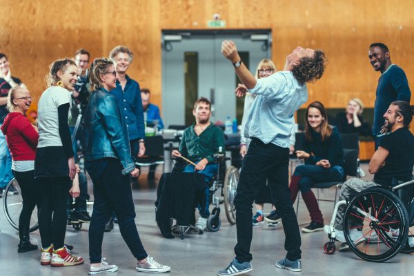 A group of people performing together in a rehearsal space. One person is sat down and 2 people are wheelchair users.