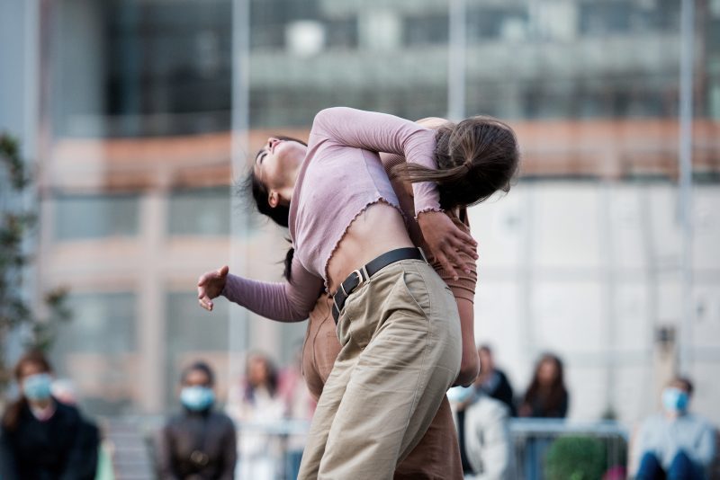 An image of 2 female dancers intertwining. The dancer facing the camera is wearing a pink cropped top and beige trousers.