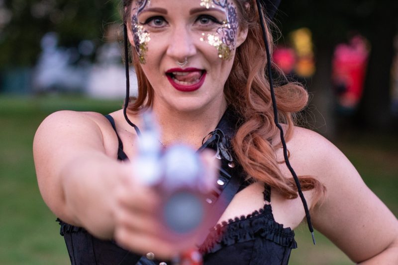 An image of a woman wearing a black hat with red hair and decorative make up facing and pointing an object at the camera.