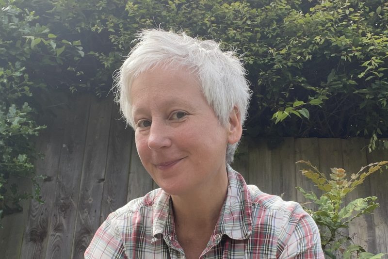 A portrait image of a woman with short grey hair facing the camera with foliage in the background.