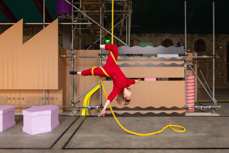 A circus performer hanging upside down from a yellow rope wearing a red boiler suit, with a sparse background.