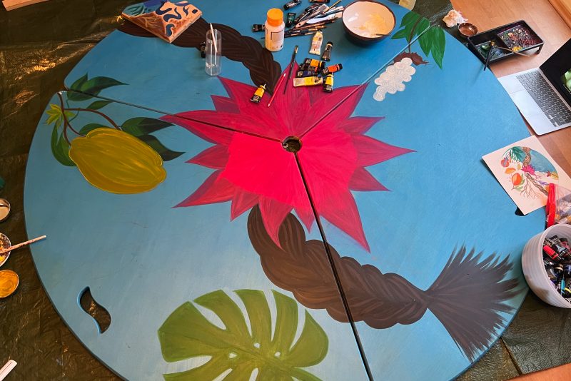 An image of a circular wooden art work decorated in blue, green and pink vibrant colours.