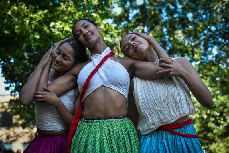 An image of three female dancers intertwining wearing traditional Indian clothing against a sunlit and leafy background.