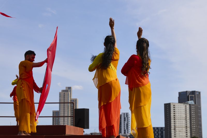 An image of three performers, 2 female and 1 male wearing traditional orange and yellow Indian dress. The 2 females have their backs to the camera and are raising their hand upwards towards the sky, and the male is holding a large red banner.