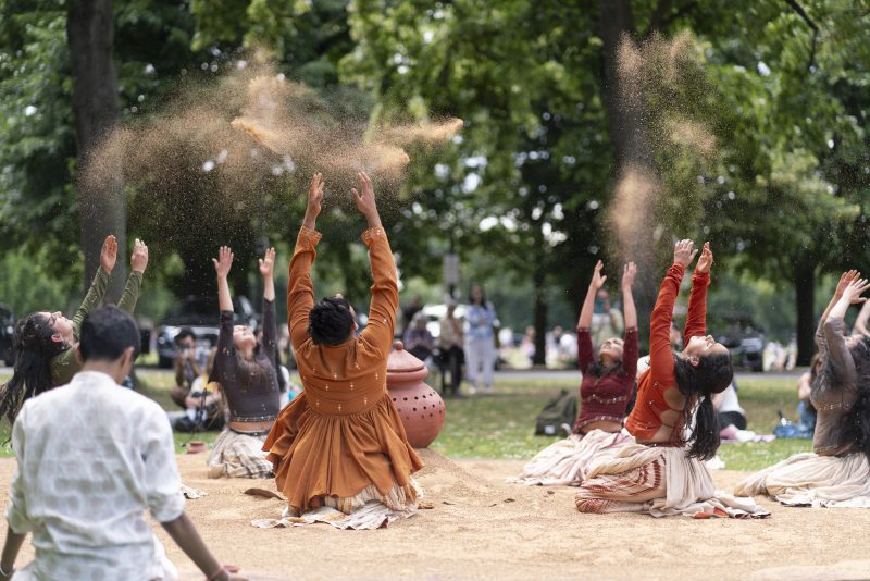 An image of a group of performers kneeling on a sandy surface. They are wearing traditional Indian dress and throwing sand into the air.