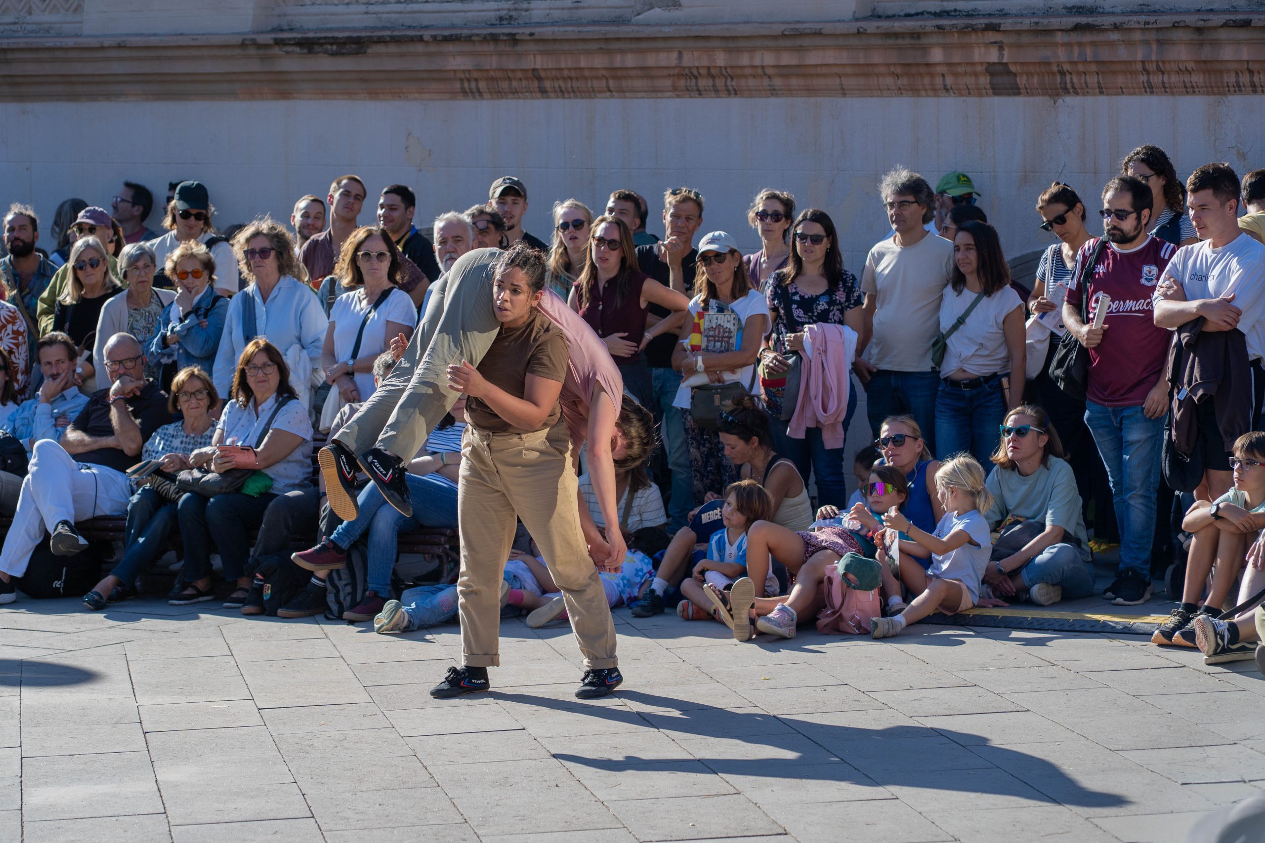 An image of 2 dancers intertwining with a crowd in the background. The dancers are casting a shadow on the concrete pavement.