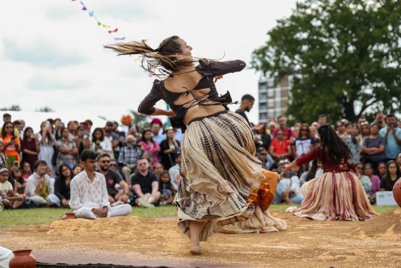 An image of a female dancer barefoot dancing on a sandy surface. She is wearing a black top and long cream and black skirt.