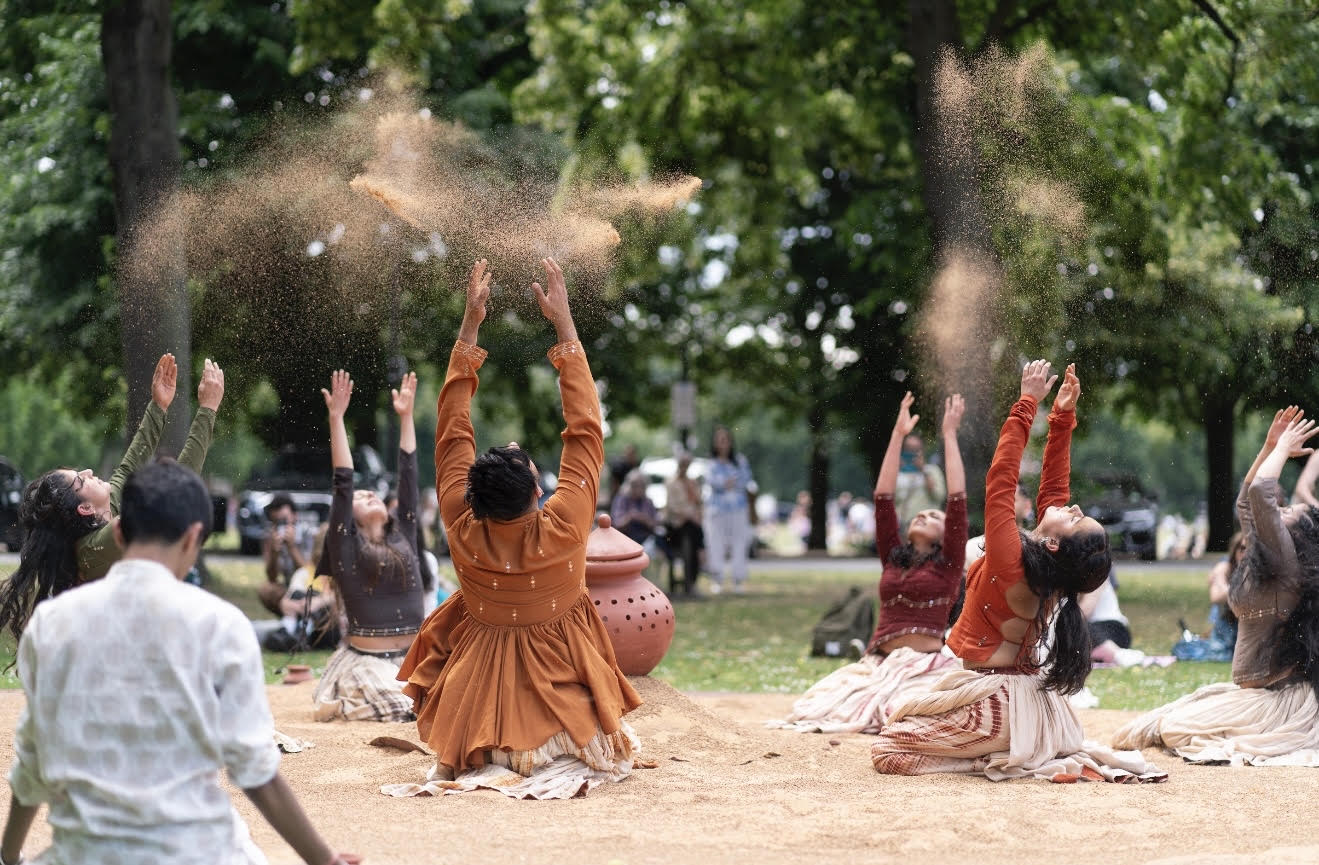 An image of a group of performers kneeling on a sandy surface wearing traditional Indian dress. They are throwing sand into the air.