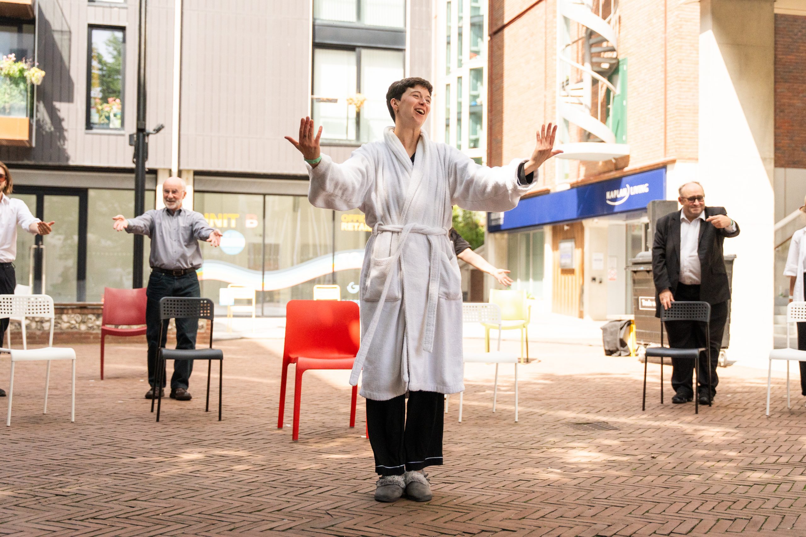 An image of a group of performers stood in front of some plastic chairs scattered around a courtyard. There is a female performer at the front of the frame wearing a white dressing gown and slippers, with her arms outstretched and she is smiling.