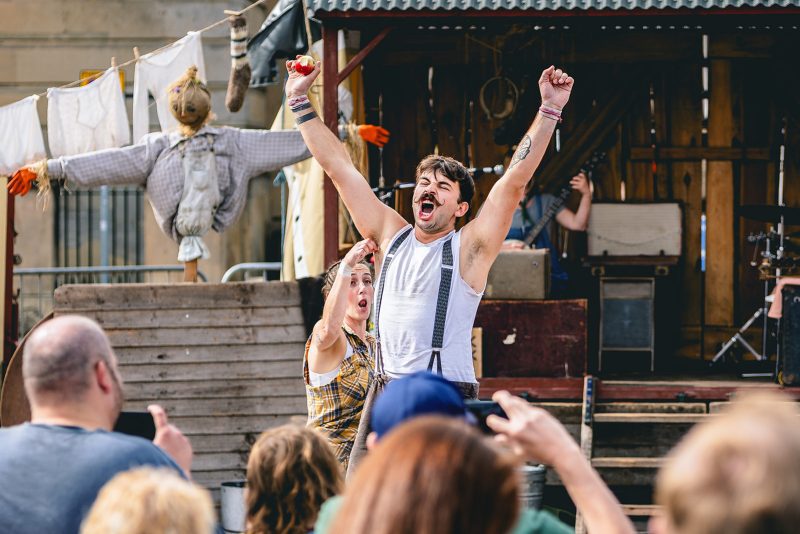 An image of a male performer with his mouth open and arms outstretched. He is wearing a white vest with braces. There is a woman placing something in his arm pit.
