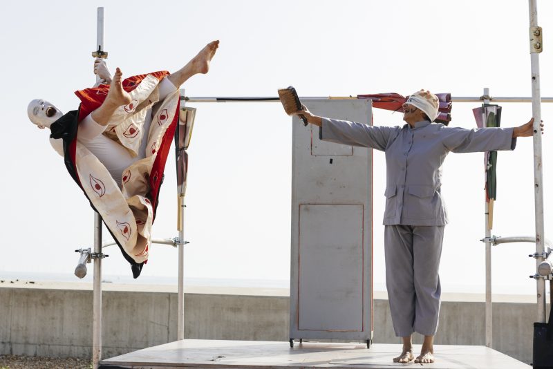 An image of a male and female performer on a makeshift platform with a sunlit sky background.